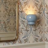 A traditional bathroom with an Ellsworth Wall Sconce 9", Blue Crackle Ceramic mounted on crane-patterned wallpaper beside a brass-framed mirror and cream wainscoting.