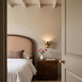 A transitional bedroom with the Stanton Wall Sconce 8", Hammered Vintage Brass mounted on the wall above a burl wood nightstand beside a striped headboard.