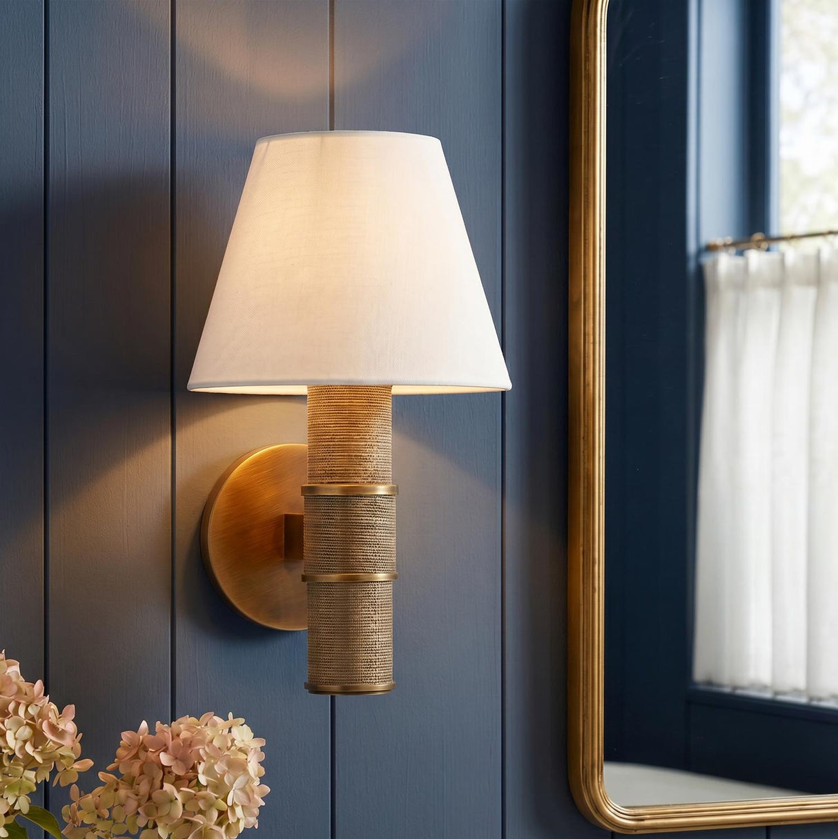 A coastal bathroom with a Hargrove Wall Sconce in patina brass and grasscloth with a white linen shade mounted on blue shiplap walls beside a gold mirror.