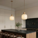 A transitional kitchen with two Norden Medium Pendant 16 inch aged brass and milk glass fixtures suspended over a black marble island and beige flat-panel cabinets.