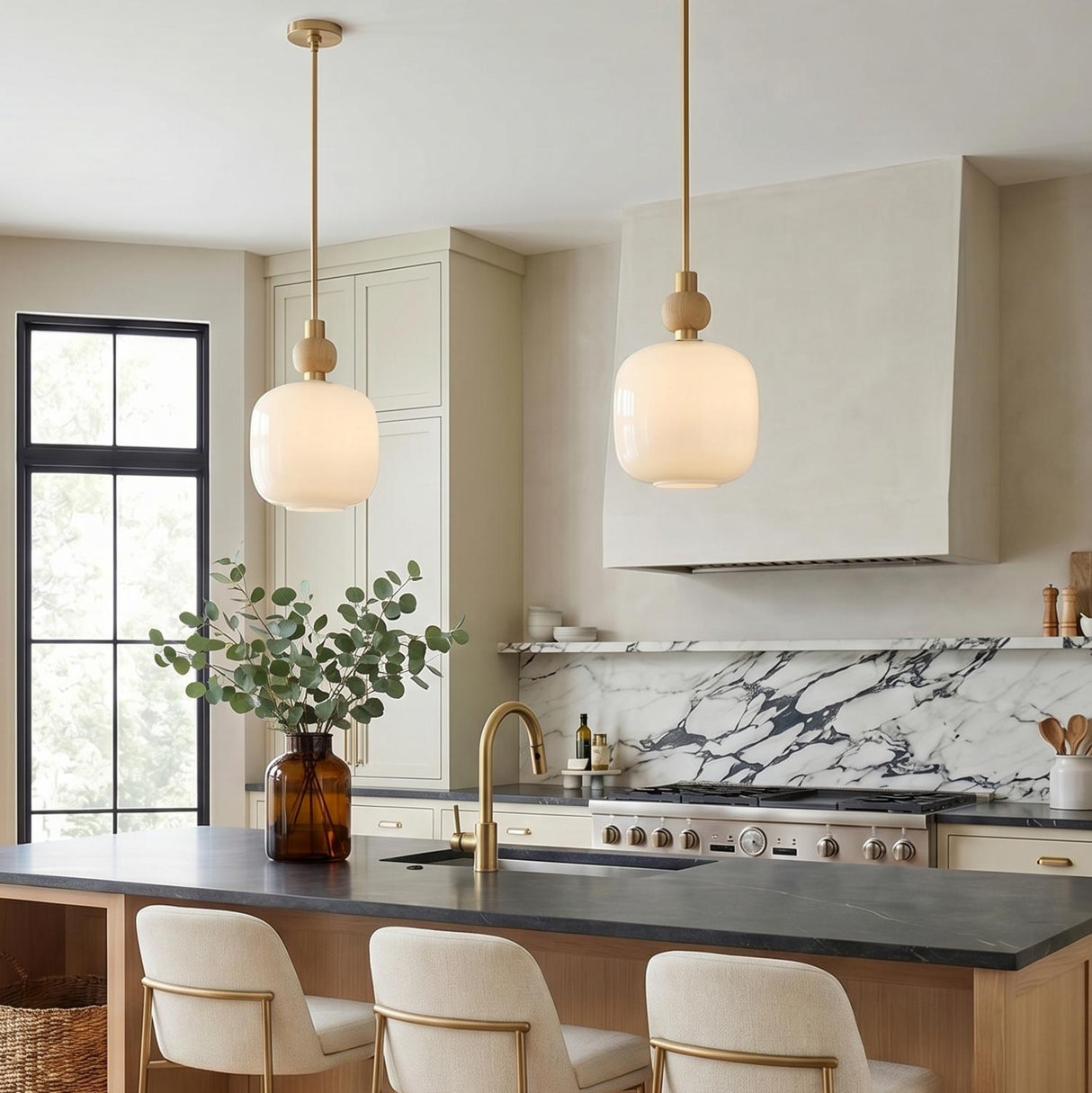 A transitional kitchen with two Norden Small Pendant, Aged Brass and Milk Glass fixtures over a black quartz island featuring a marble backsplash and beige cabinets.