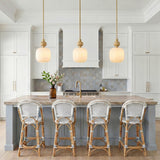 A coastal kitchen with three Norden Small Pendants in aged brass and milk glass over a stone-topped island with woven rattan stools and coffered ceilings.