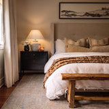 A transitional bedroom featuring the Kensington Table Lamp 24", Sand Ceramic and Aged Brass on a black nightstand beside a beige upholstered headboard and wooden bench.