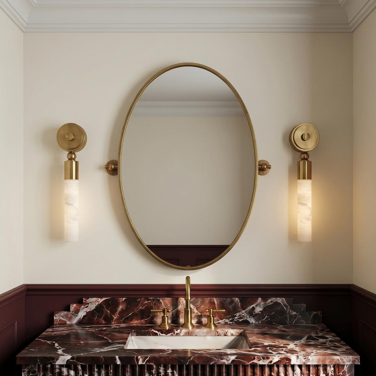 A transitional bathroom with two Arden LED Wall Sconces in vintage brass and alabaster flanking an oval mirror above a dark red marble vanity with burgundy wainscoting.
