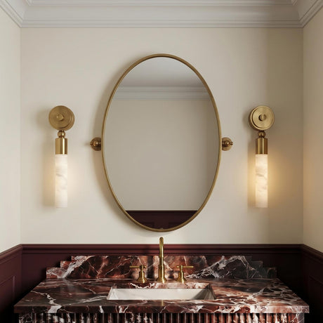 A transitional bathroom with two Arden LED Wall Sconces in vintage brass and alabaster flanking an oval mirror above a dark red marble vanity with burgundy wainscoting.