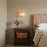 A traditional bedroom with the Bristolia Wall Sconce in aged brass and clear piastra glass mounted above a scalloped wood nightstand featuring a woven wicker basket.
