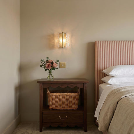 A traditional bedroom with the Bristolia Wall Sconce in aged brass and clear piastra glass mounted above a scalloped wood nightstand featuring a woven wicker basket.