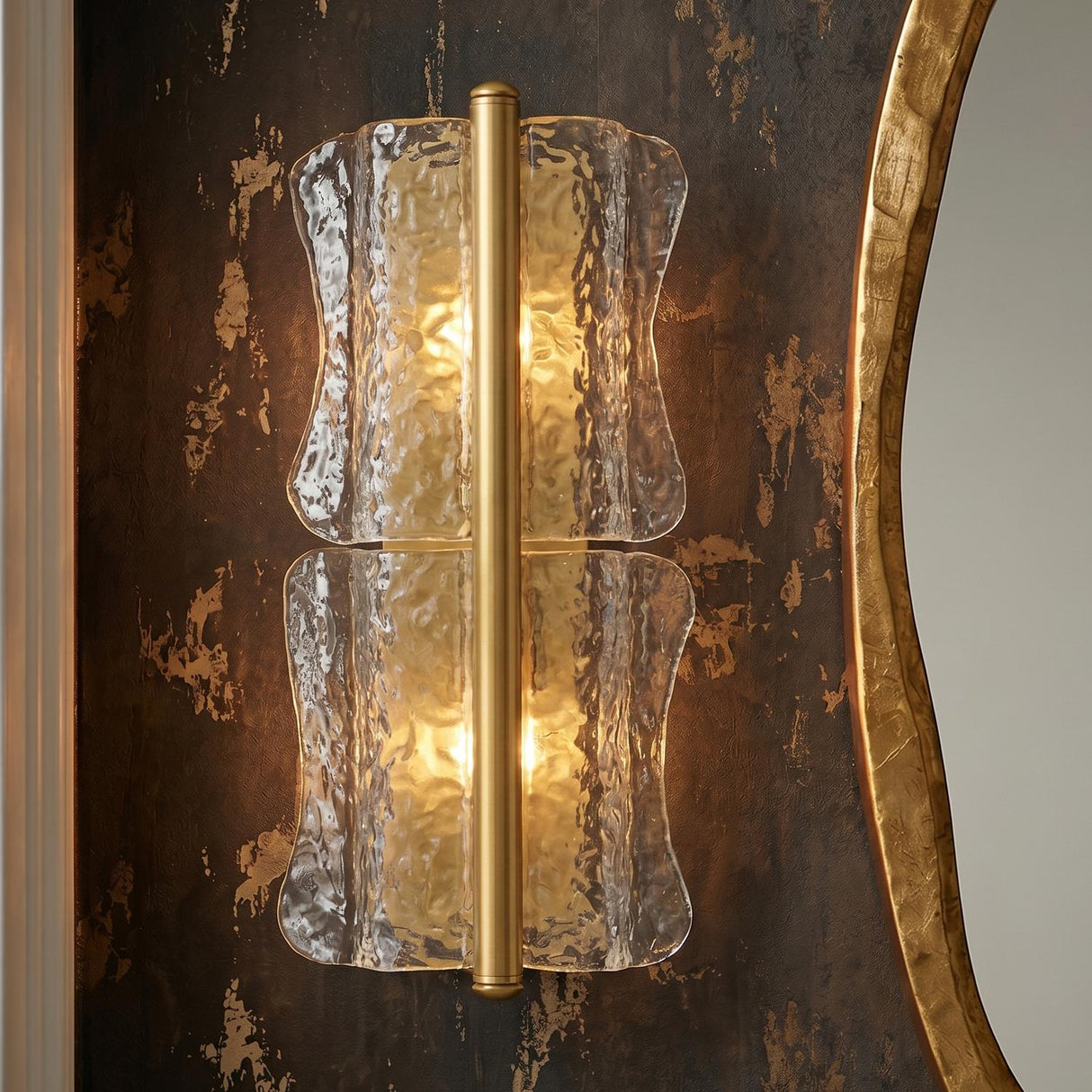 A contemporary bathroom featuring the Bristolia 2 Light Wall Sconce in aged brass and clear piastra glass mounted on a dark textured wall beside a gold mirror.