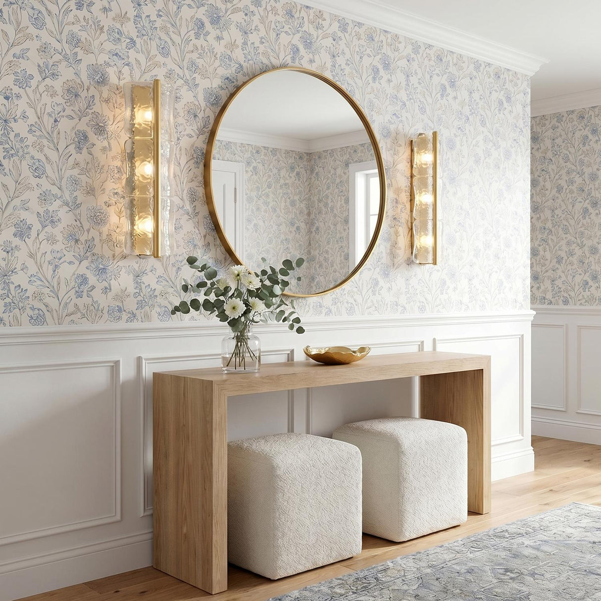 A transitional foyer with two aged brass and clear piastra glass wall sconces flanking a round gold mirror above a wood console table and floral wallpaper.