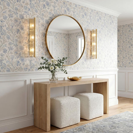 A transitional foyer with two aged brass and clear piastra glass wall sconces flanking a round gold mirror above a wood console table and floral wallpaper.