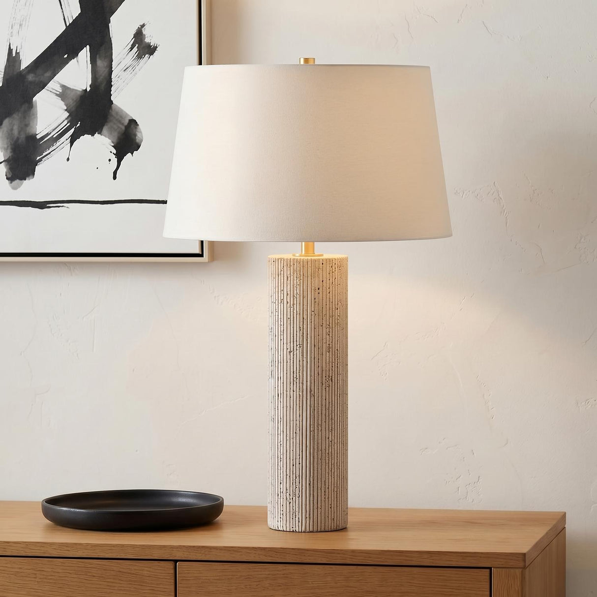 A minimalist living room with a Cassian Table Lamp 30 inch featuring a fluted travertine base and linen shade on a wood console beside black abstract art.