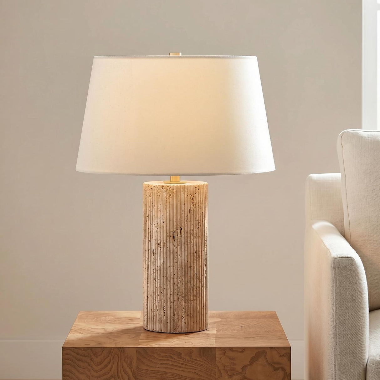 A contemporary living room with the Cassian Table Lamp featuring a fluted travertine base and linen shade on a wood side table beside a cream sofa.