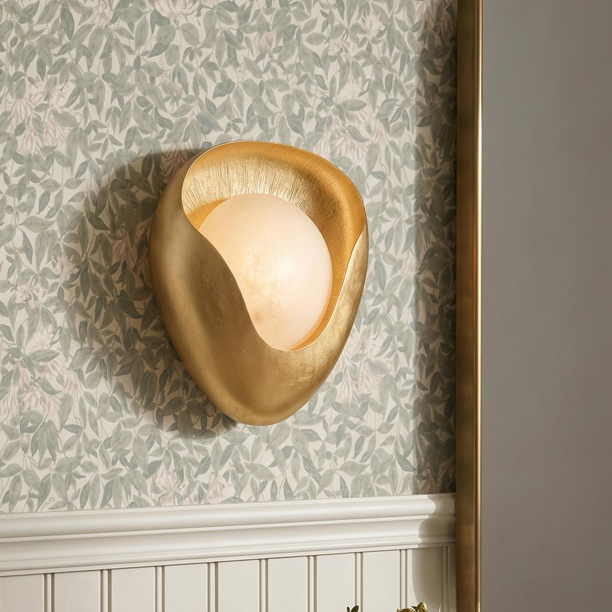A transitional foyer with a Jovie LED Wall Sconce in textured gold leaf and alabaster mounted against floral wallpaper above white beadboard wainscoting.