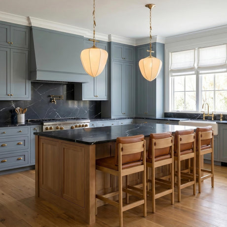 A transitional kitchen with two Chiya Medium Pendants in vintage brass and white silk over a black marble island with blue cabinetry and wood floors.