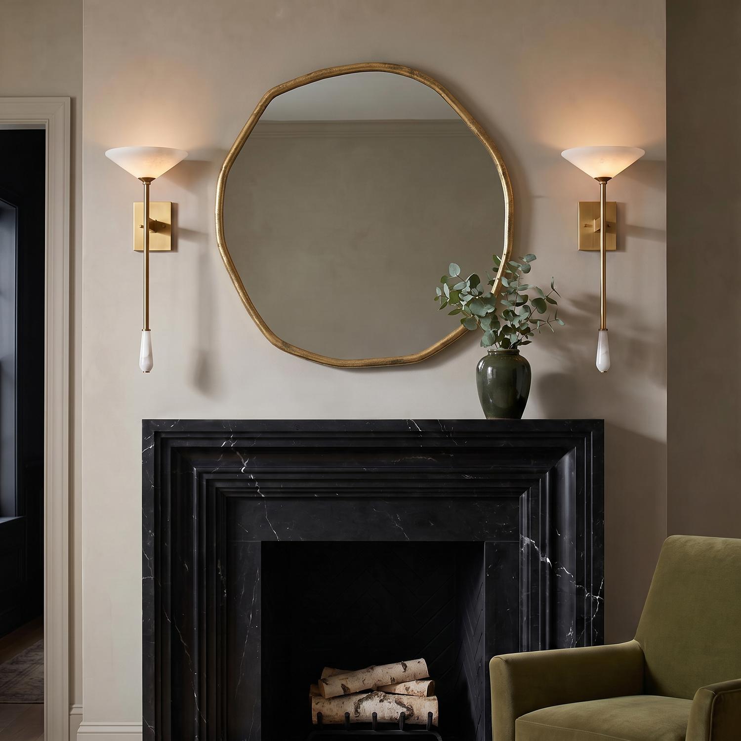 A contemporary living room with two Seneca LED wall sconces in vintage brass and alabaster flanking an organic gold mirror above a black marble fireplace.