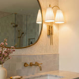 A transitional bathroom featuring the Wynn Double Wall Sconce 24", Vintage Brass with pleated linen shades mounted beside a round mirror over a marble vanity and grey tile.