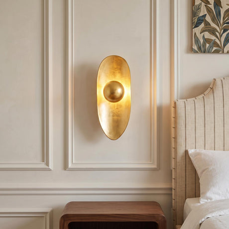 A transitional bedroom featuring a Syon LED Wall Sconce 19", Gold Leaf mounted on a white paneled wall beside a striped fabric headboard and walnut nightstand.