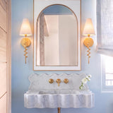 A transitional bathroom features two Linnea Wall Sconce 21", Gold Leaf fixtures with fabric shades flanking an arched mirror above a scalloped marble vanity.