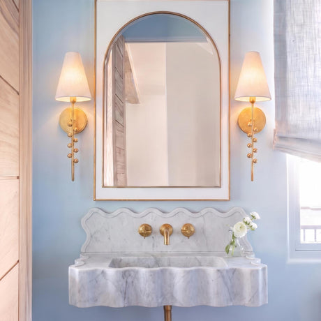 A transitional bathroom features two Linnea Wall Sconce 21", Gold Leaf fixtures with fabric shades flanking an arched mirror above a scalloped marble vanity.