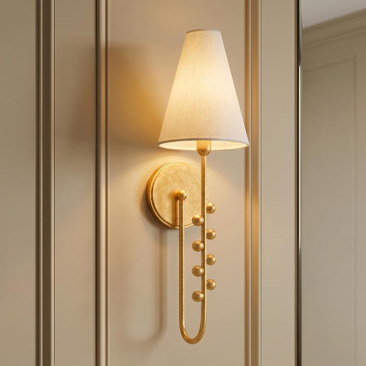 A traditional foyer with the Linnea Wall Sconce in a gold leaf finish featuring a white linen shade and beaded detailing on beige wall paneling.