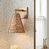 A coastal bathroom featuring a Grove Wall Sconce with braided jute and patina brass mounted on blue leaf-patterned wallpaper beside a gold vanity mirror.