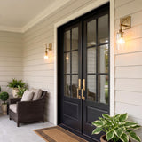A modern farmhouse porch featuring the Webster Outdoor Wall Light in patina brass flanking black double doors on beige siding with a wicker armchair and potted greenery.