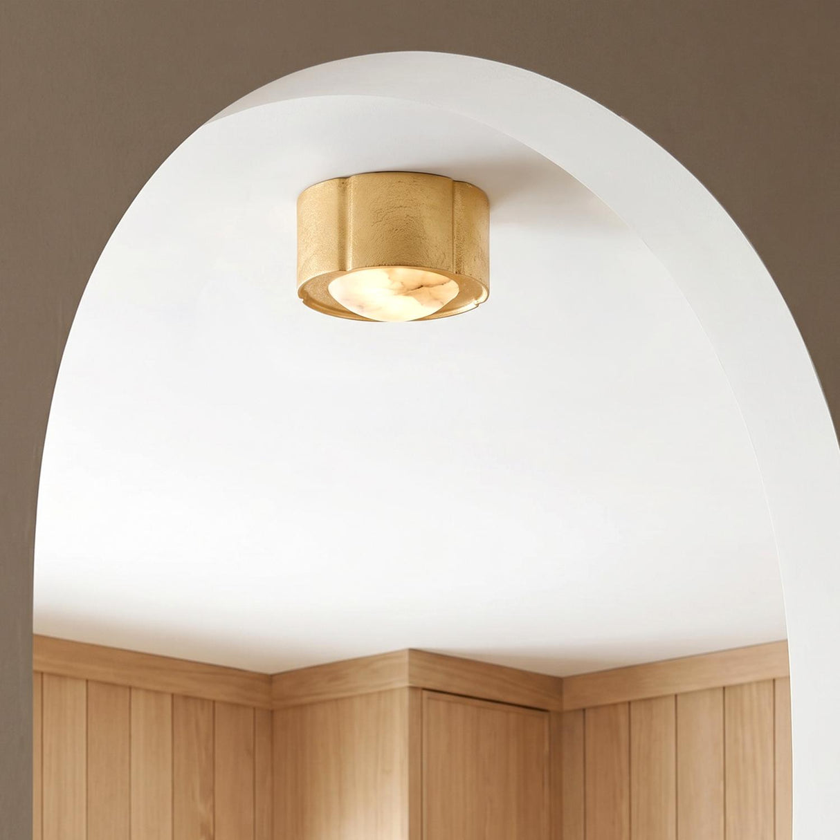 A transitional foyer with a gold leaf and alabaster flush mount installed on a white ceiling within a deep archway above light wood wall paneling.