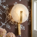 A traditional bedroom with the Zahara LED Wall Sconce in Alabaster and Gold Leaf mounted on dark floral wallpaper beside a gold-framed mirror and pink hydrangeas.