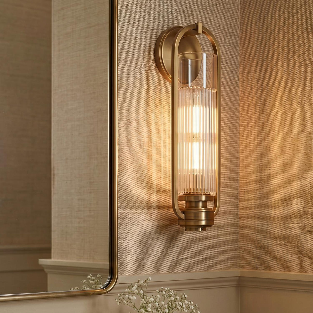 A contemporary bathroom with an aged brass wall sconce featuring ribbed glass and an oval frame, mounted beside a thin-framed mirror on beige grasscloth wallpaper.