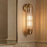 A contemporary bathroom with an aged brass wall sconce featuring ribbed glass and an oval frame, mounted beside a thin-framed mirror on beige grasscloth wallpaper.