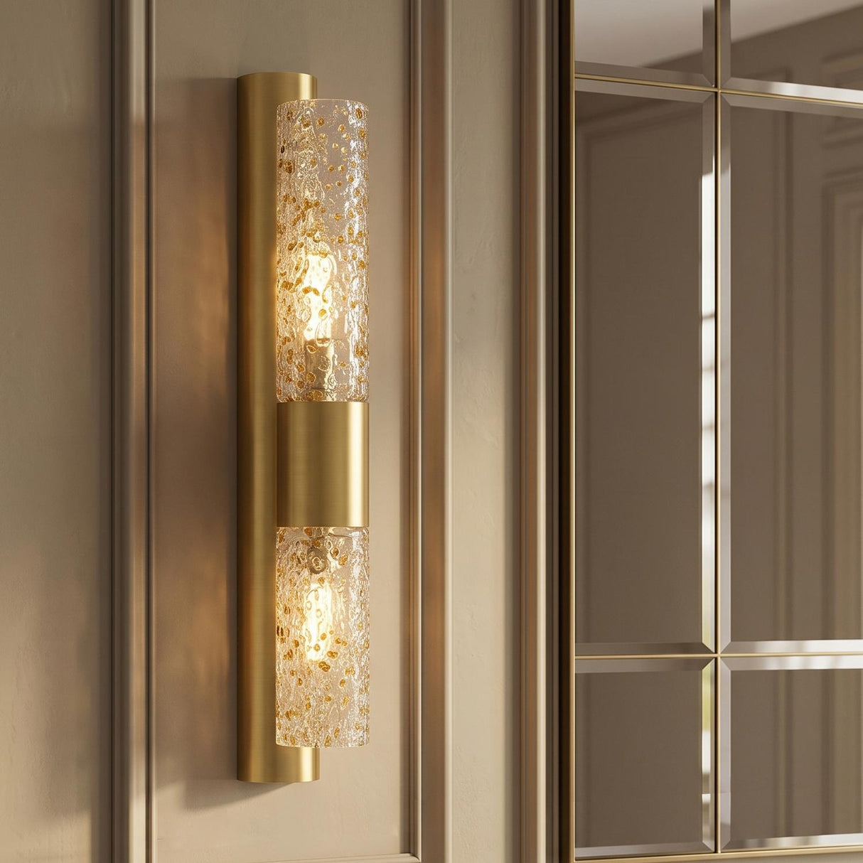 A transitional foyer with the Daylin Wall Sconce 24", Vintage Brass and Amber Speckled Glass mounted on beige paneled walls beside a large gold grid mirror.