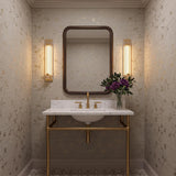 A transitional bathroom with two gold leaf and reeded glass wall sconces flanking a dark wood mirror above a marble console vanity with tan floral wallpaper.