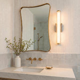 A contemporary bathroom with a Gaven LED Wall Sconce in gray travertine and aged brass mounted beside an organic mirror above a white marble vanity with brass faucets.
