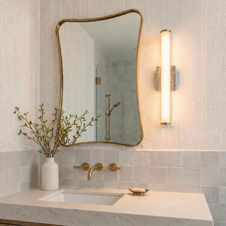 A contemporary bathroom with a Gaven LED Wall Sconce in gray travertine and aged brass mounted beside an organic mirror above a white marble vanity with brass faucets.