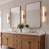 A transitional bathroom with three Gaven LED wall sconces in gray travertine and aged brass flanking two mirrors over a white marble double vanity and wood cabinetry.