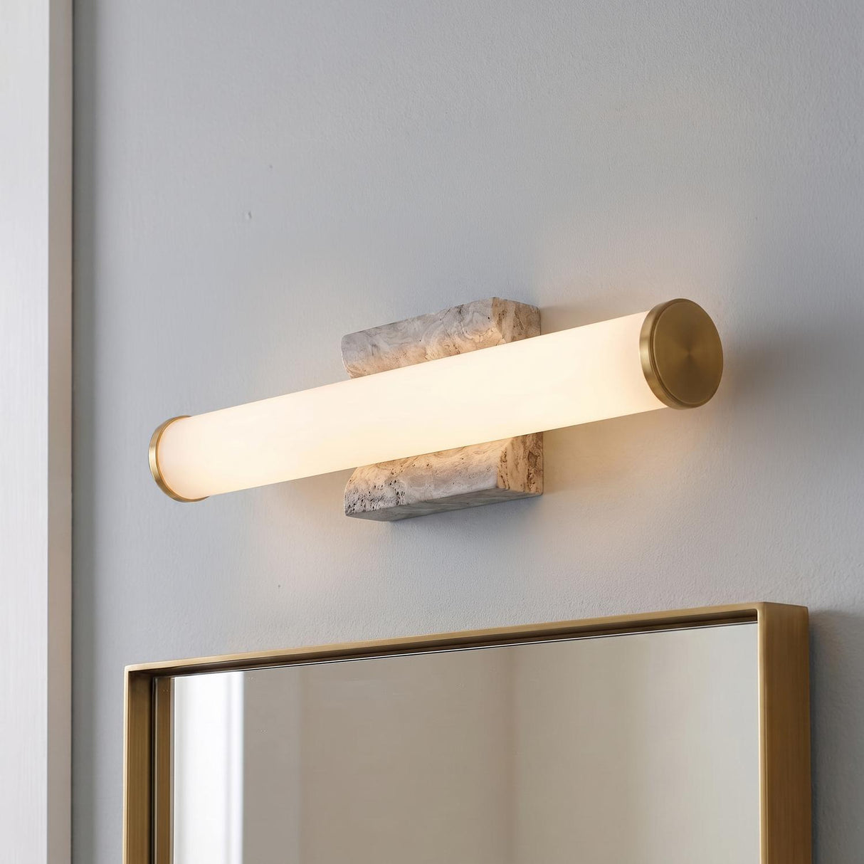 A modern bathroom with a gray travertine and aged brass Gaven LED Wall Sconce 25" mounted above a matching brass-framed mirror on a neutral wall.