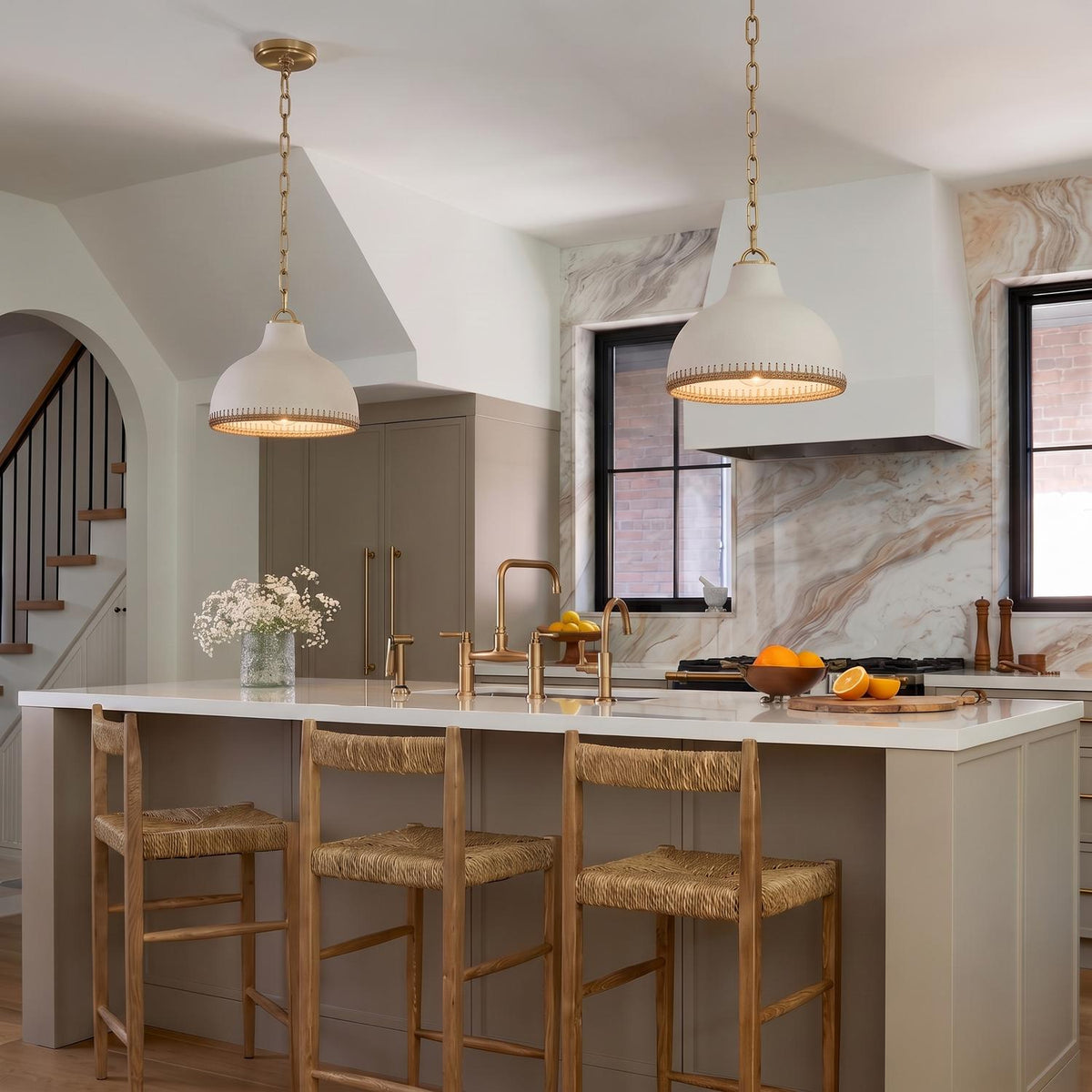 A transitional kitchen with two Nadira Medium Pendants in cream and rattan with aged brass hardware hanging over a white quartz island beside a marble backsplash.