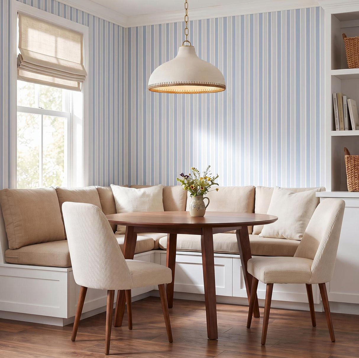 A cottage dining room with a cream and rattan pendant in aged brass over a round wood table, built-in banquette, and blue striped wallpaper.