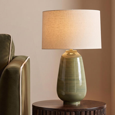 A transitional living room with the Prunella Table Lamp featuring a glossy olive ceramic base and wheat linen shade on a dark wood table beside a green sofa.