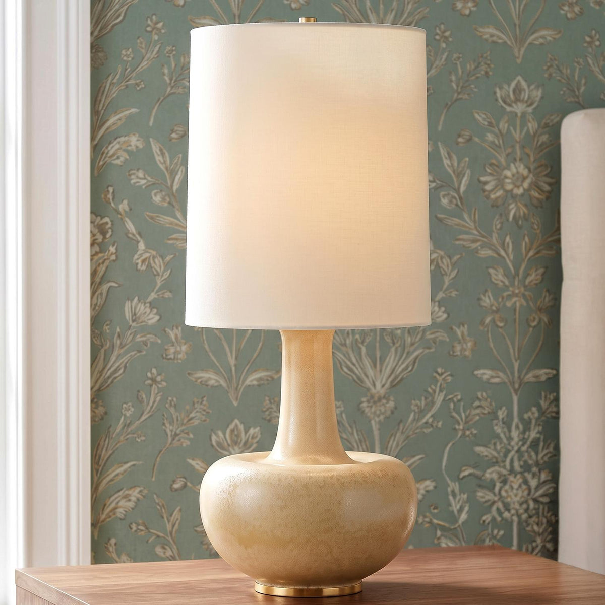 A traditional bedroom featuring the Viviana Table Lamp 28", Biscotti Glaze with a white linen drum shade on a wood nightstand beside green floral wallpaper.