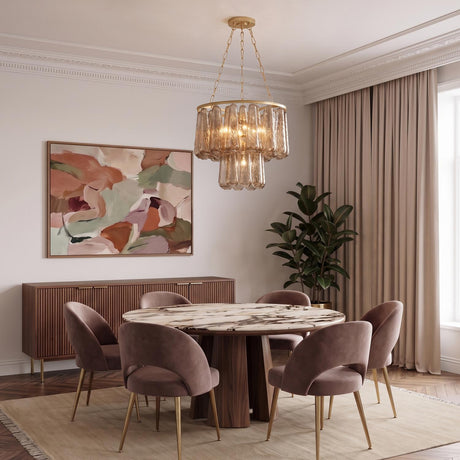 A contemporary dining room featuring a gold leaf chandelier with smoke taupe swirled glass over a round marble table, mauve velvet chairs, and a slatted wood sideboard.