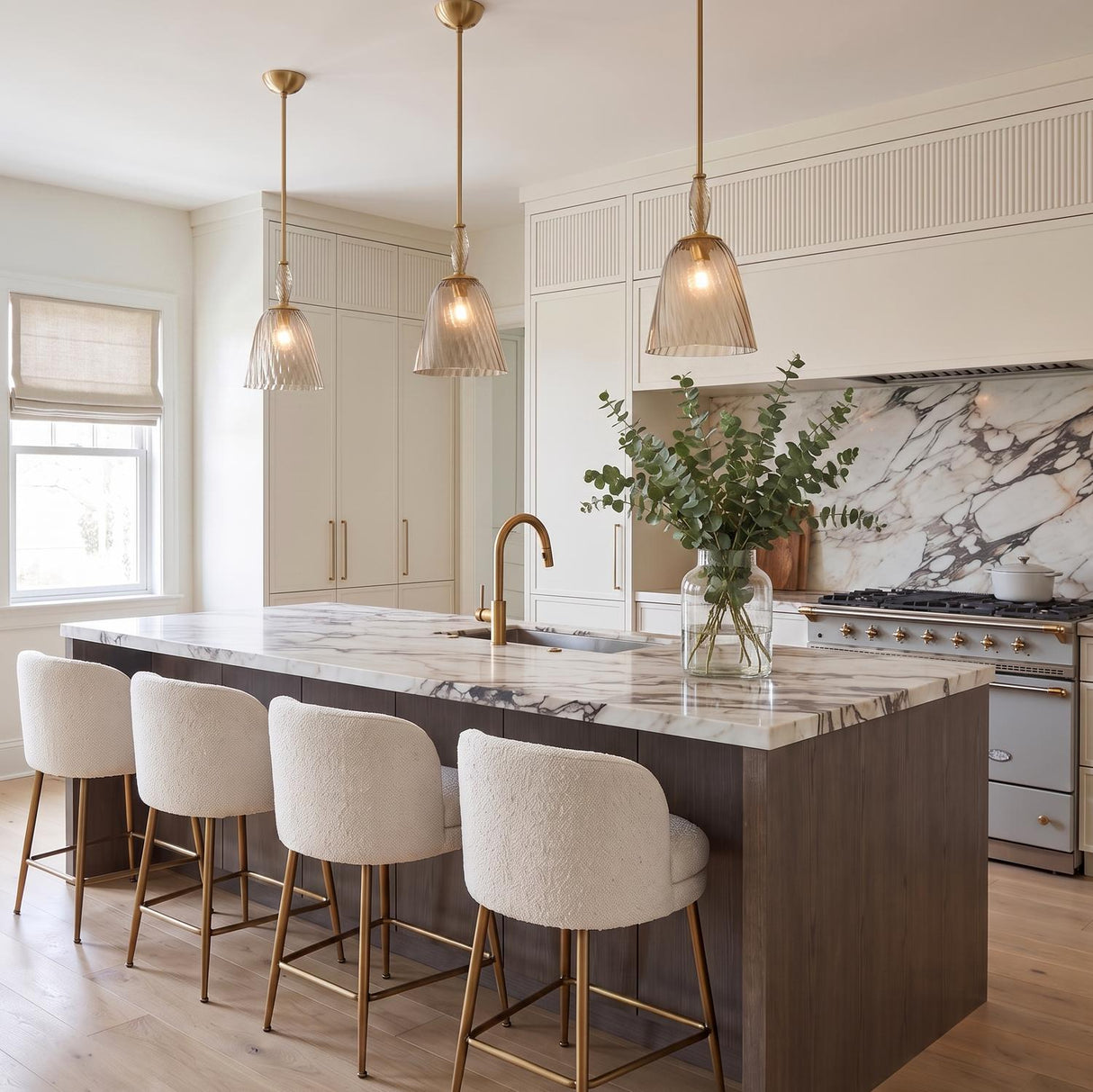 A transitional kitchen featuring three Bly Small Pendants in aged brass and smoke taupe glass over a marble island with bouclé stools and marble backsplash.
