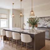 A transitional kitchen featuring three Bly Small Pendants in aged brass and smoke taupe glass over a marble island with bouclé stools and marble backsplash.