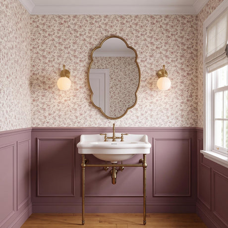A traditional bathroom with two patina brass and fluted milk glass wall sconces flanking a scalloped mirror above a white console sink and mauve floral wallpaper.