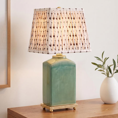 A transitional living room featuring a jade glaze ceramic table lamp with a pleated primrose printed shade placed on a wooden console table beside a white vase.