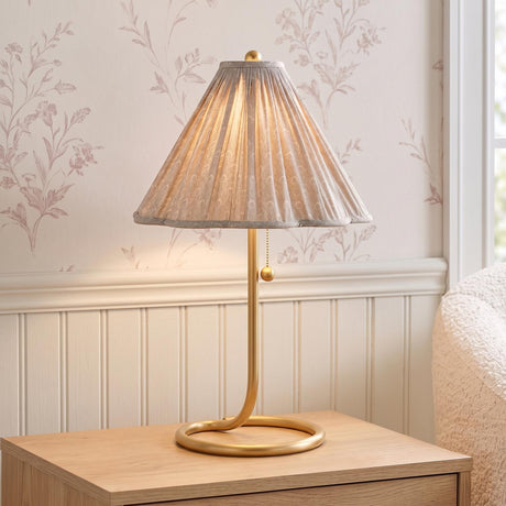 A traditional bedroom featuring the Nina Table Lamp in aged brass and a taupe printed shade on a light wood nightstand against floral wallpaper and beadboard.
