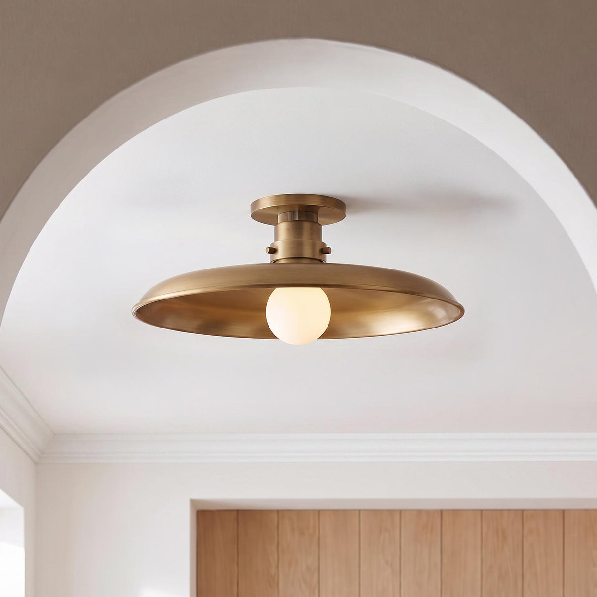 A transitional foyer with the Titan Semi-Flush Mount 16 inch in Patina Brass and frosted glass globe, centered on a white ceiling archway above wood paneling.