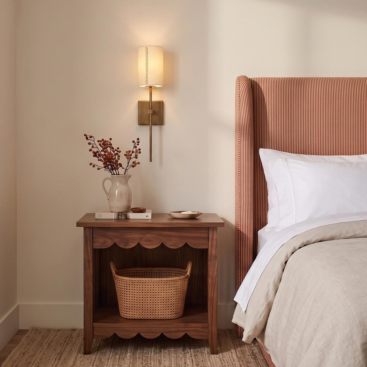 A transitional bedroom with a patina brass wall sconce and linen shade mounted above a scalloped walnut nightstand beside a pink pinstripe upholstered headboard.