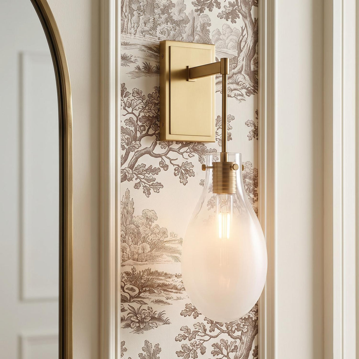 A transitional bathroom with a Geneva Wall Sconce 19", Brass and Ombre Frosted Glass mounted beside an arched brass mirror on brown toile wallpaper.