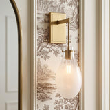 A transitional bathroom with a Geneva Wall Sconce 19", Brass and Ombre Frosted Glass mounted beside an arched brass mirror on brown toile wallpaper.